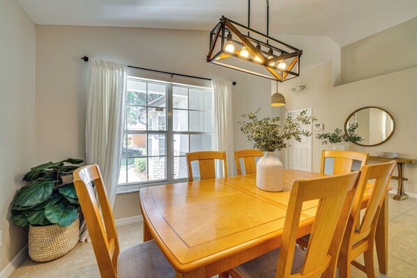 Un área de comedor con una mesa y sillas de madera, una lámpara colgante, un ventanal con cortinas y una planta en maceta.