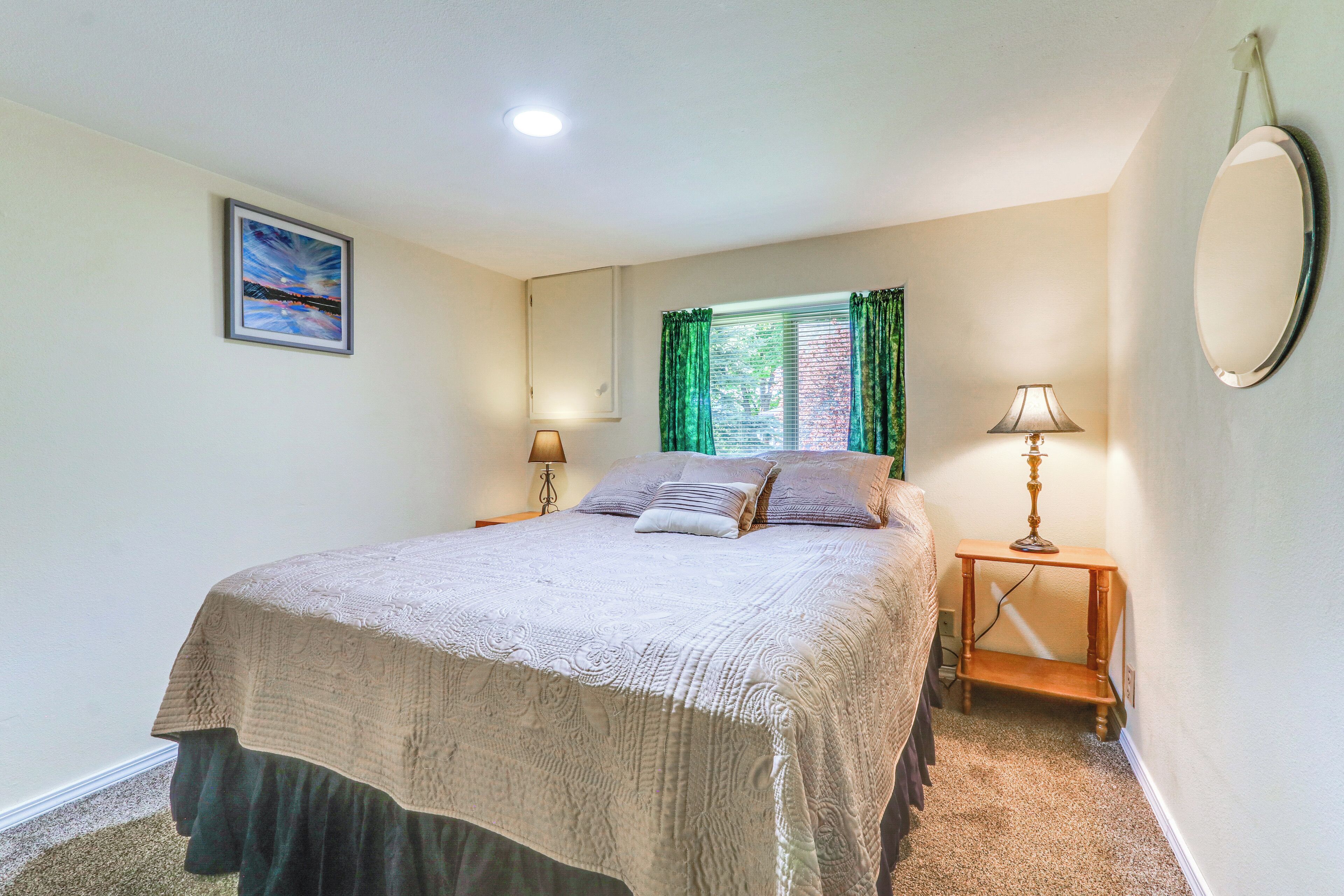 A bedroom with a bed, two bedside tables, a window with curtains, a wall-mounted mirror, and a painting on the wall.