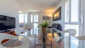 A modern living room with a glass dining table, white chairs, a flat-screen TV, and a potted plant.