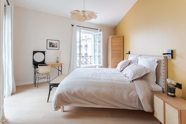 A bedroom with a bed, a desk, a chair, a wardrobe, and a window with a view of buildings.