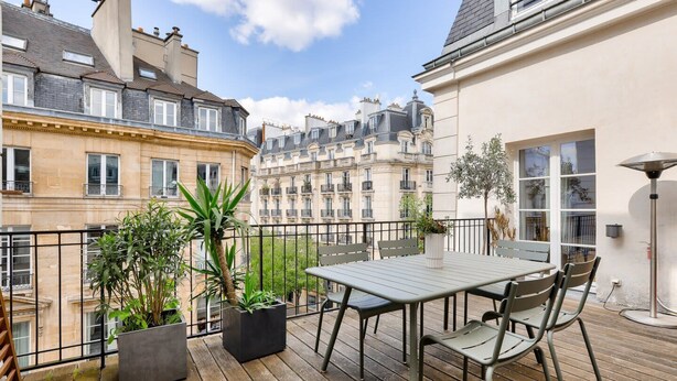 A wooden deck with a table and chairs, surrounded by potted plants and overlooking a row of classic European buildings. - Large terrace, air conditioning, 3 bedrooms, Marais (Paris)