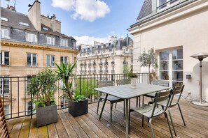 A wooden deck with a table and chairs, surrounded by potted plants and overlooking a row of classic European buildings.
