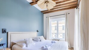 A bedroom with a large bed, a wooden headboard, a window with curtains, and a wooden nightstand with a water bottle. - Large terrace, air conditioning, 3 bedrooms, Marais (Paris)