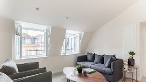 A modern living room with a grey sofa, a wooden coffee table, and a view of buildings through large windows.