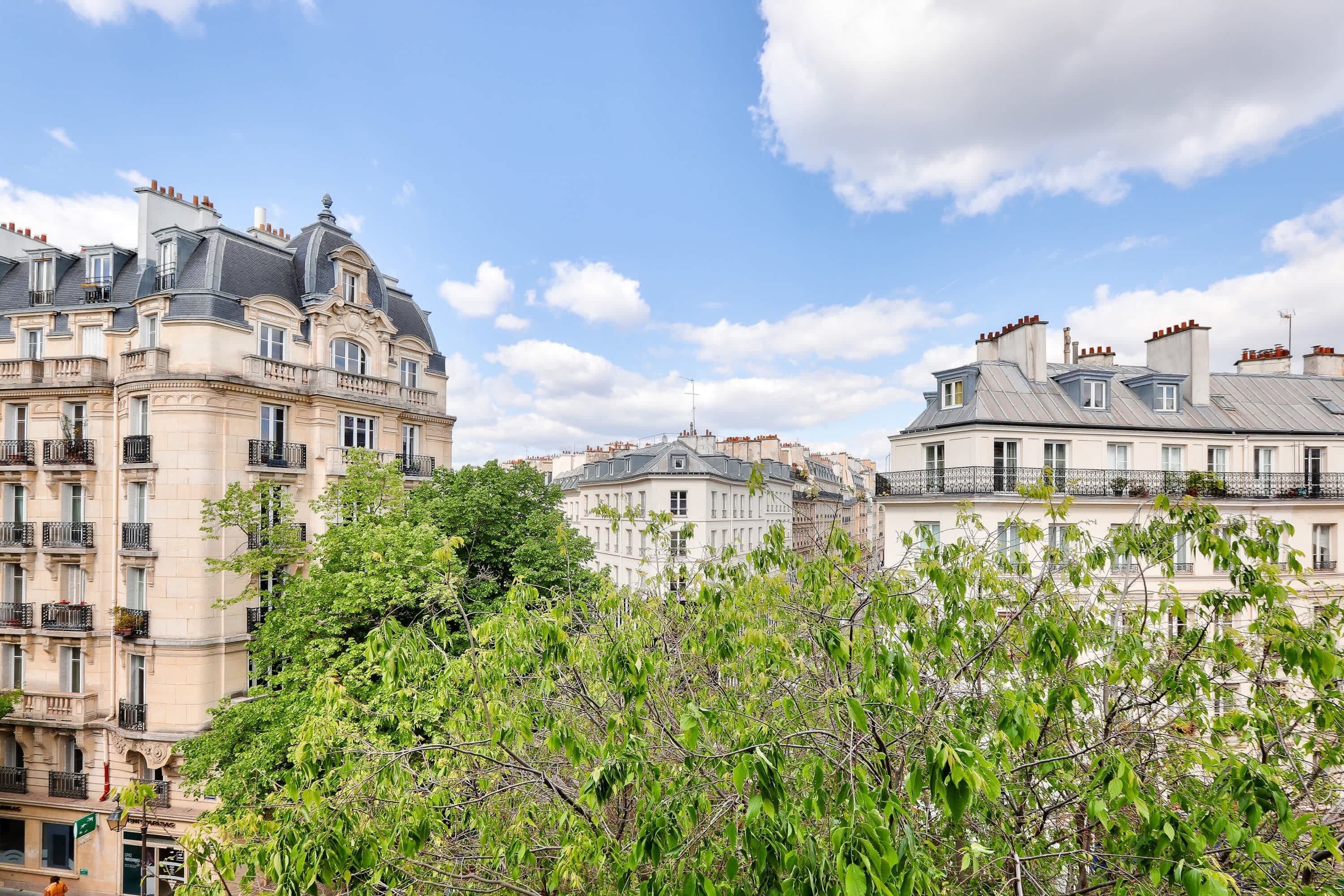 A row of historic buildings with balconies, a clear sky, and lush green trees in the foreground.