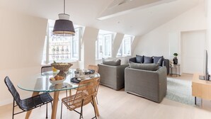 A modern living room with a glass dining table, grey sofas, and a large window.