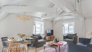 A modern loft with exposed wooden beams, a glass dining table, grey sofas, and a flat-screen TV. - Charming Apartment with Air Conditioning - Marais (Paris)
