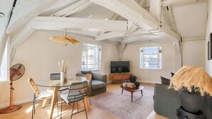 A spacious loft with exposed wooden beams, a glass dining table, a sofa, a television, and a large window. - Charming Apartment with Air Conditioning - Marais (Paris)