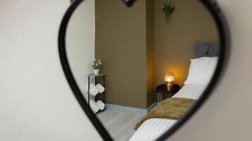 A heart-shaped mirror reflecting a bedroom with a bed, a nightstand, a lamp, and a plant.