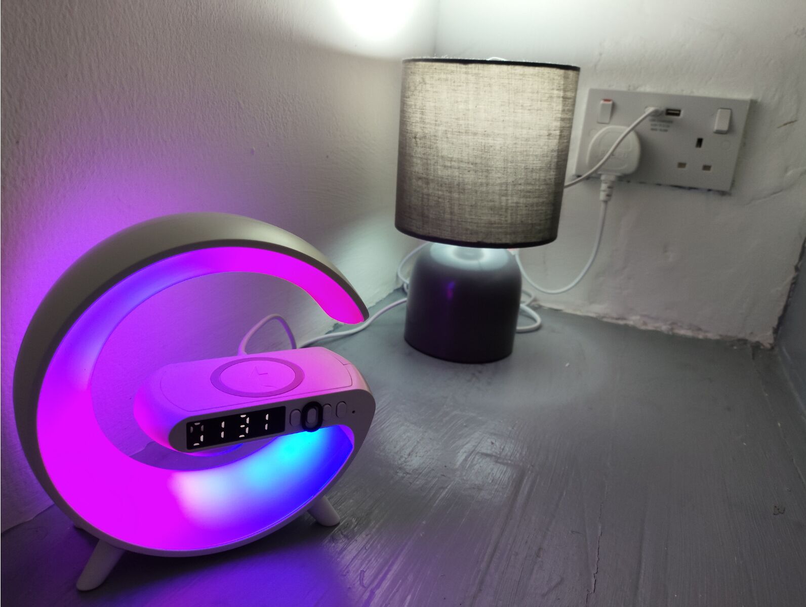 A modern, illuminated clock with a digital display and a lamp with a fabric shade on a wooden surface.
