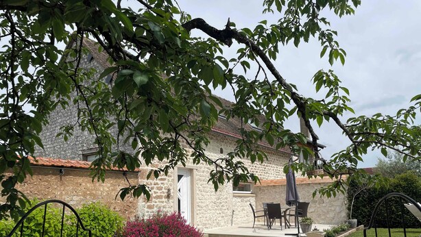 En bygning i sten med et tag af teglsten, en terrasse med udendørs møbler og en grussti.