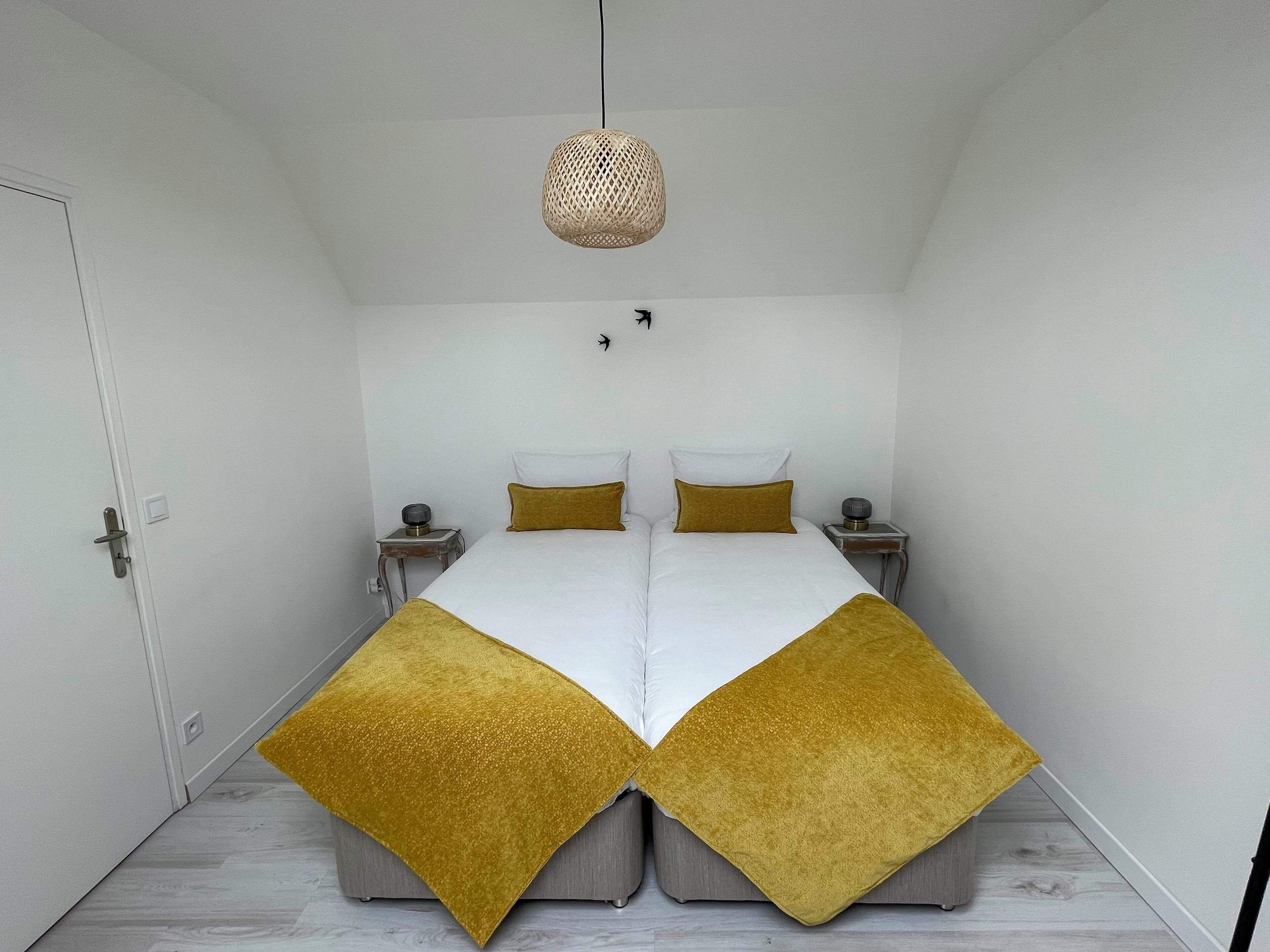 Cama doble con dos almohadas blancas y dos mantas doradas, una mesita de noche con una lámpara, y una lámpara de techo tejida.