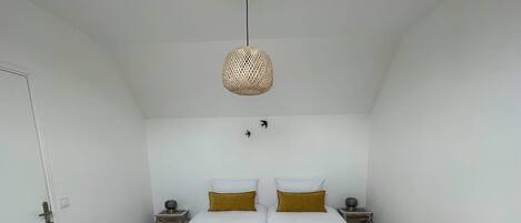 Cama doble con dos almohadas blancas y dos mantas doradas, una mesita de noche con una lámpara, y una lámpara de techo tejida.
