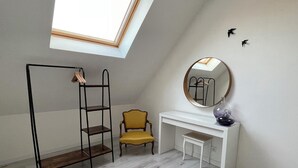 Et værelse med ovenlys, et hvidt skrivebord med et spejl, en gul stol og en sort metalreol.