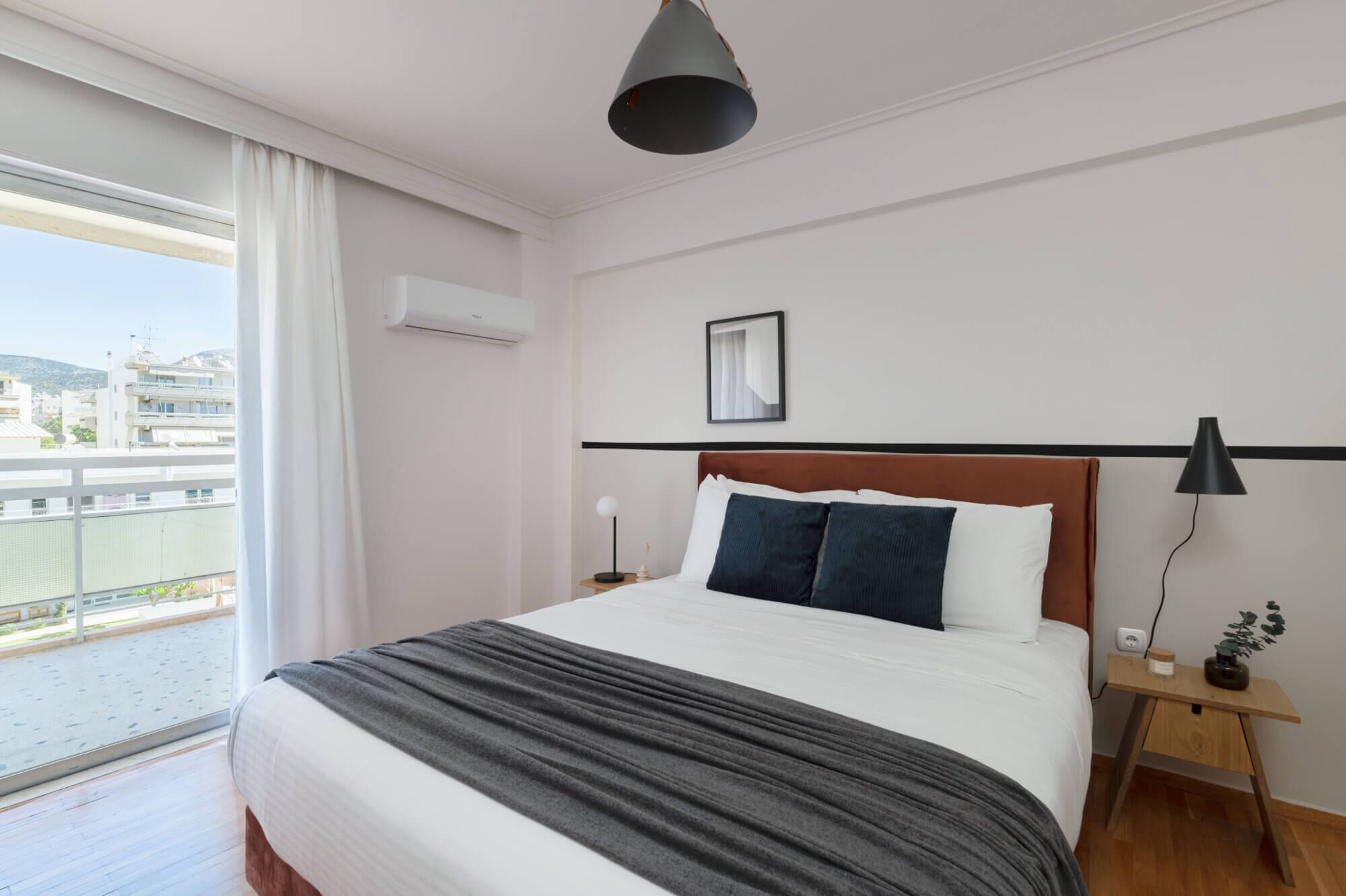 Une chambre d’hôtel avec un grand lit, une table de chevet, une commode, une lampe et une vue sur la ville par la fenêtre.