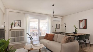 Een moderne woonkamer met een bank, een salontafel en een eethoek met een tafel en stoelen. Er is een radiator, een plant en een plafondlamp.