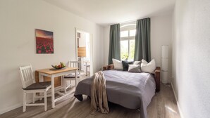 Ein Schlafzimmer mit Bett, Nachttisch und Schreibtisch mit Stuhl. Es gibt ein Fenster mit Vorhängen, ein Bild an der Wand und eine Lampe neben dem Bett.