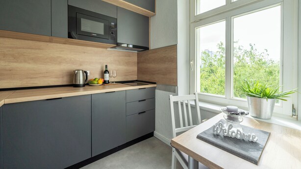 Eine moderne Küche mit grauen Schränken, einer Holz Arbeitsplatte, einer eingebauten Mikrowelle, einem Wasserkocher, einer Weinflasche und einem Topfpflanzen am Fenster.