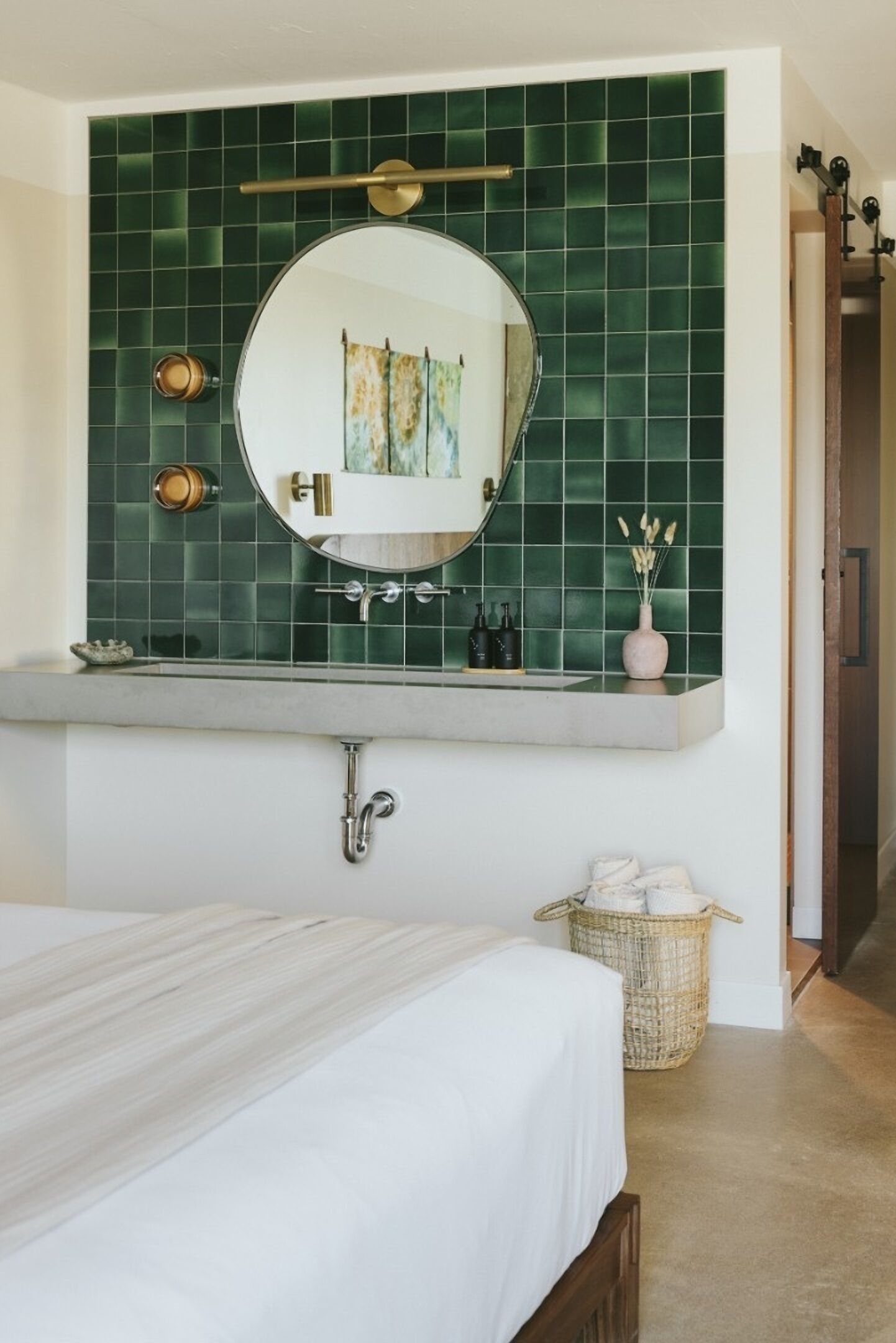 Een badkamer met een groen betegelde muur, een ronde spiegel, een handdoekrek en een rieten mand.