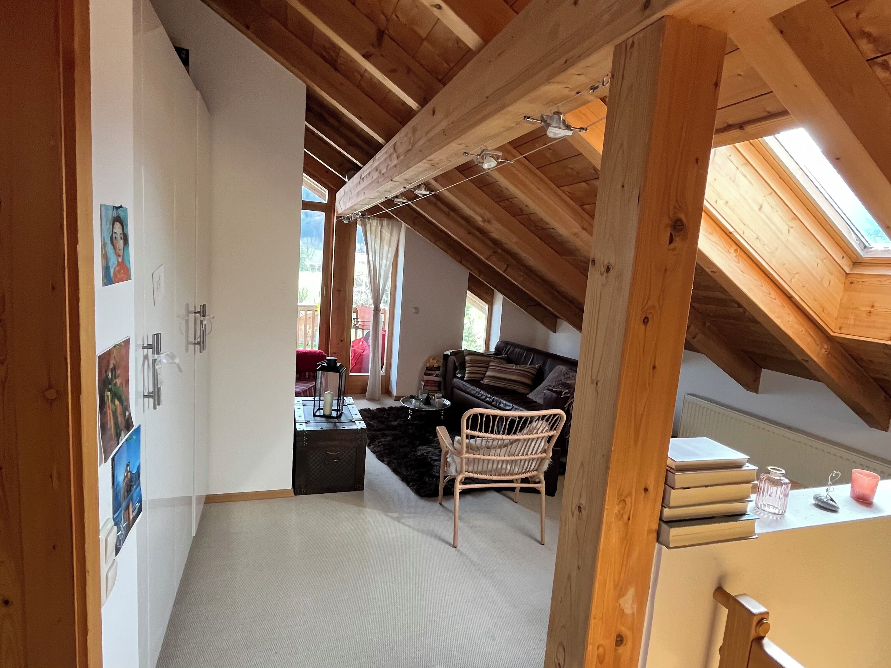 Una habitación acogedora en el ático, con techo inclinado, vigas de madera, una silla de mimbre, un sofá y una mesita con libros y una vela.
