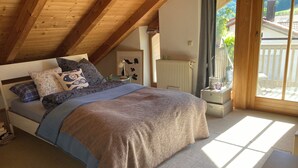 A bedroom with a sloped ceiling, a bed with a blue and brown bedspread, a nightstand with decorative items, and a view of the outdoors through a large window. - Ferienwohnung Kampenwinkel 68 qm, Schlafzimmer, Wohn-/schlafzimmer, Südbalkon (Aschau im Chiemgau)