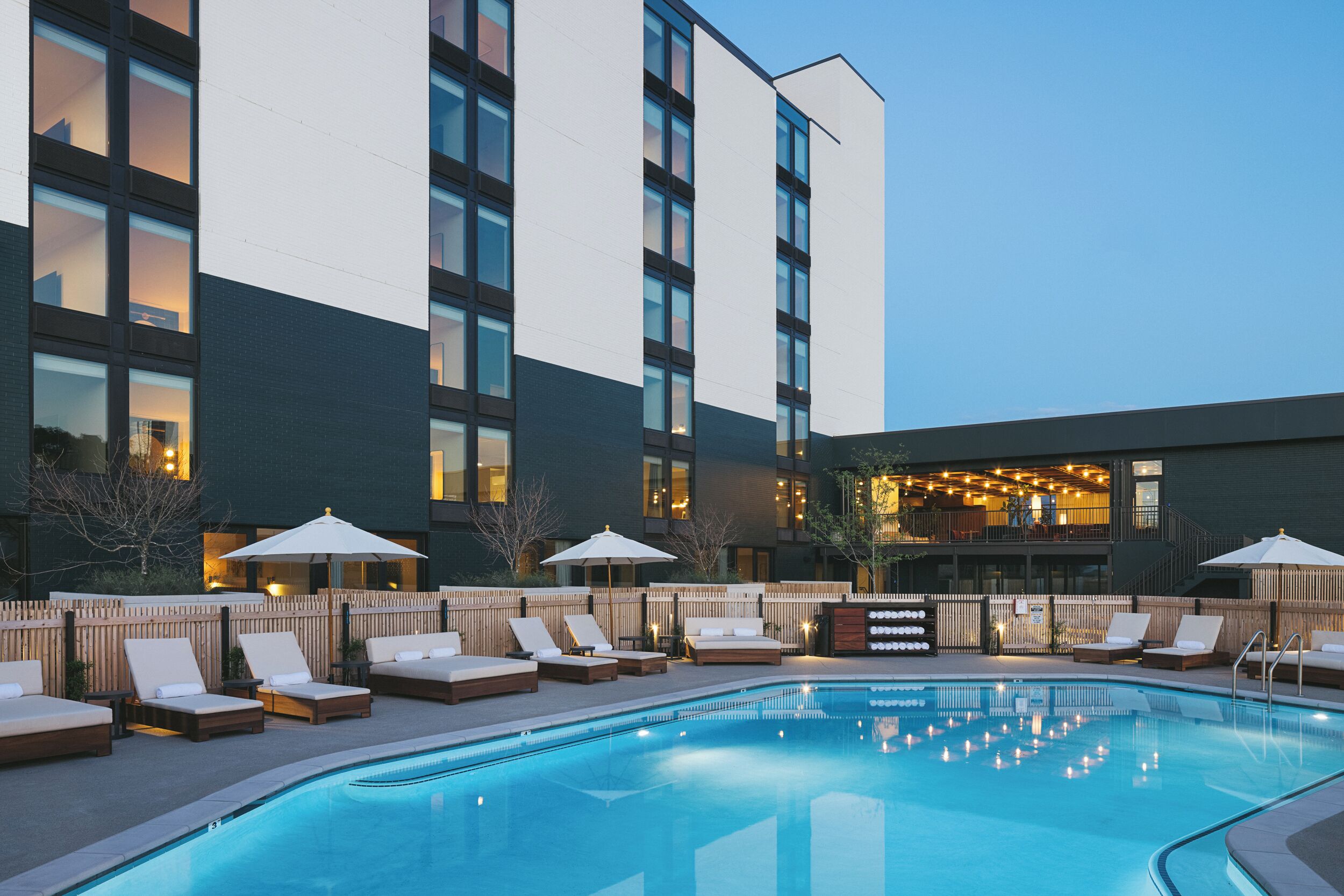 A pool area with lounge chairs and umbrellas, a bar with lights, and a building with multiple floors and windows.