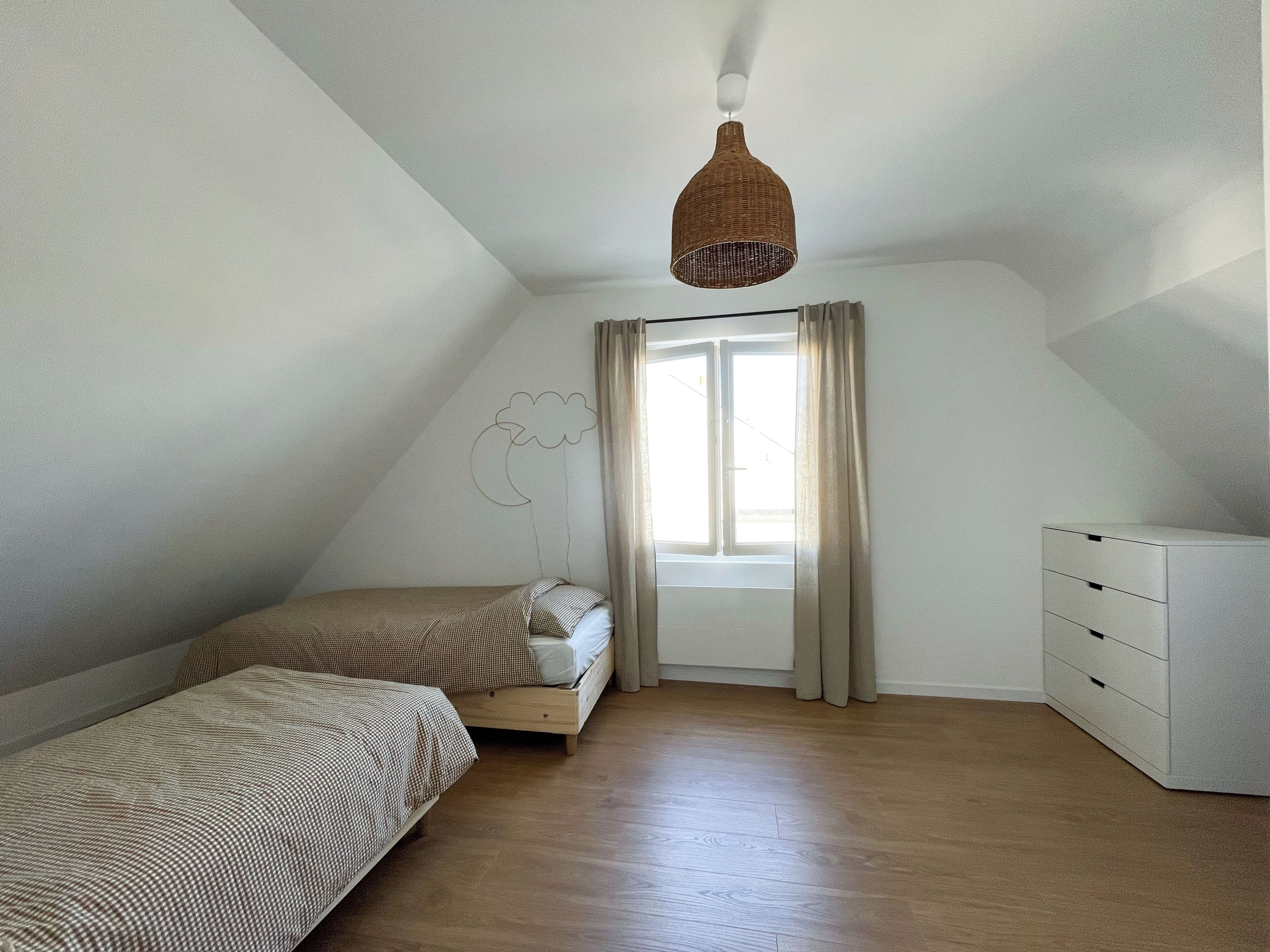 A bedroom with two single beds, a wooden dresser, a hanging light fixture, a window with curtains, and a wall-mounted light fixture.