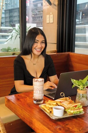 A person sitting at a wooden table with a laptop, a drink, and a tray of food including a sandwich and fries. - Connector Hostel (Makati)