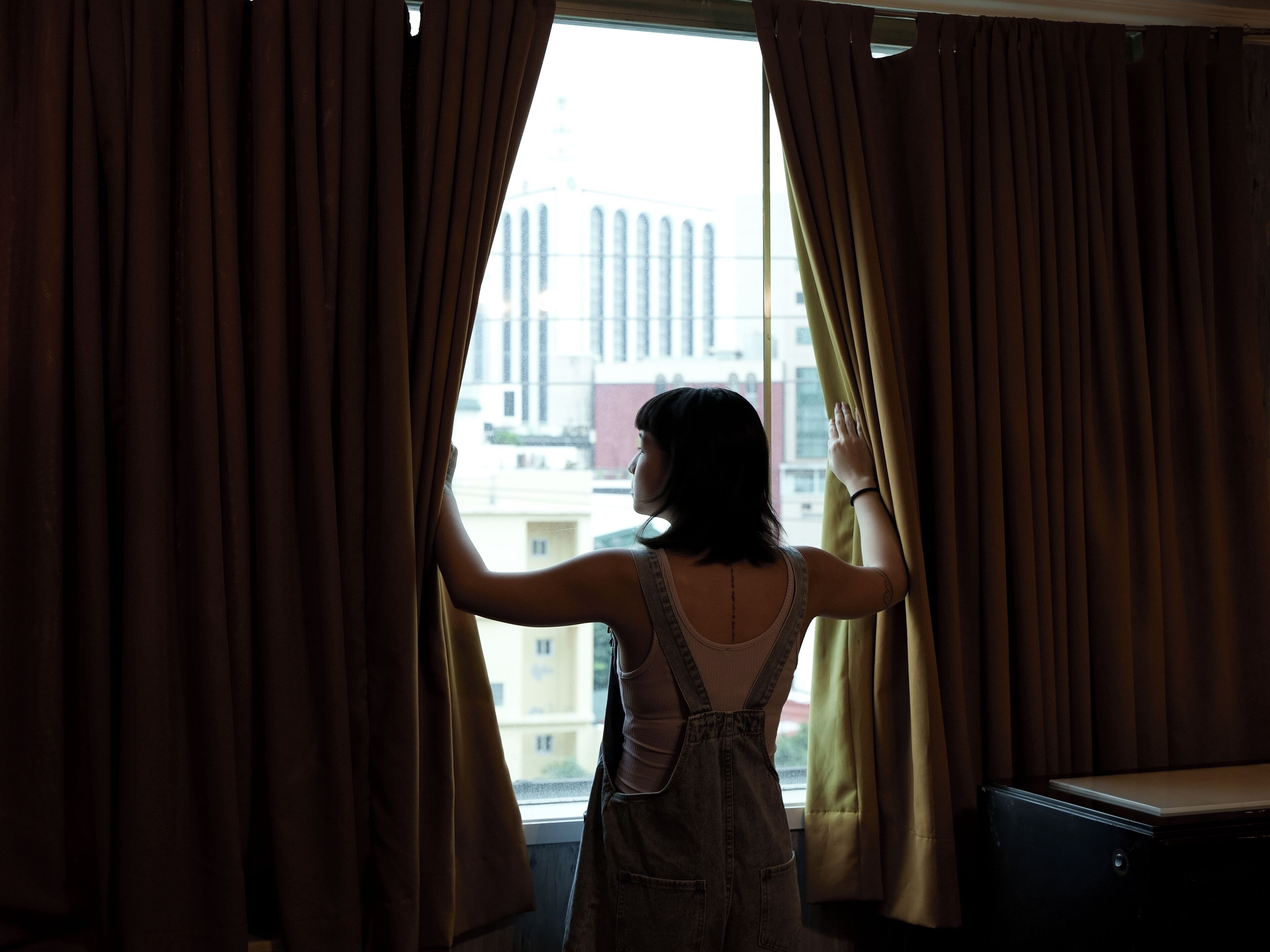 A person standing by a window with curtains, looking outside.
