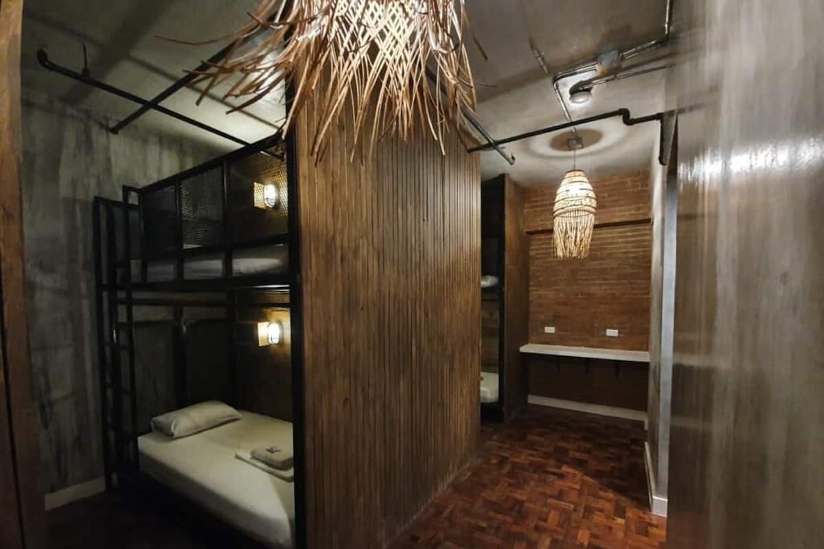 A compact hotel room with a bunk bed, a wooden column, a hanging light fixture, and a small bathroom area.