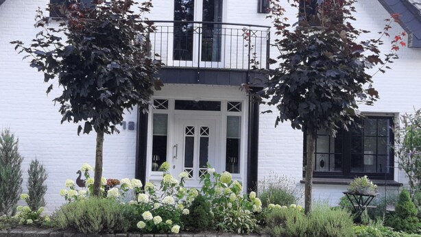 Ein weißes Haus mit Balkon, einer Haustür und einem kleinen Garten mit weißen Blüten.