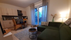 Ein kompakter Wohnraum mit einem grünen Sofa, einem Essbereich mit Stühlen, einer Küche mit Schränken und einem großen Fenster mit Vorhängen.