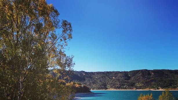 A beach with clear blue water, sandy shore, and trees with autumn leaves. - Maison de Campagne Tilleul (Beynes)