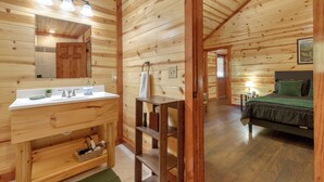 A wooden cabin interior with a bathroom featuring a sink, mirror, and shelves. A bed with a green blanket is visible in the adjacent room.
