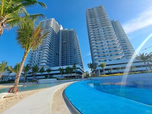 Un hotel moderno con piscina, palmeras y edificios altos al fondo.