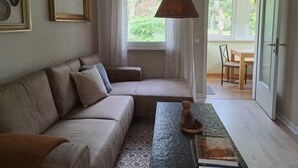 Een woonkamer met een bank, een bijzettafeltje met decoratieve objecten, een raam met gordijnen en een deur die naar een andere kamer leidt.
