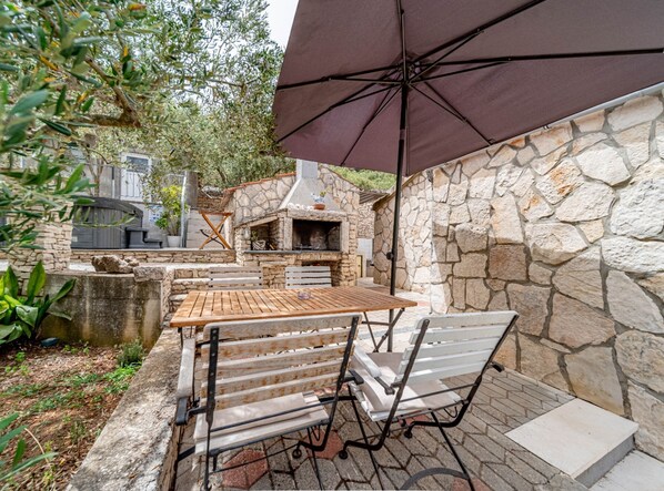 An outdoor patio with a stone wall, a wooden table, two metal chairs, and a large umbrella. - Stone House Blaca (Smokvica)