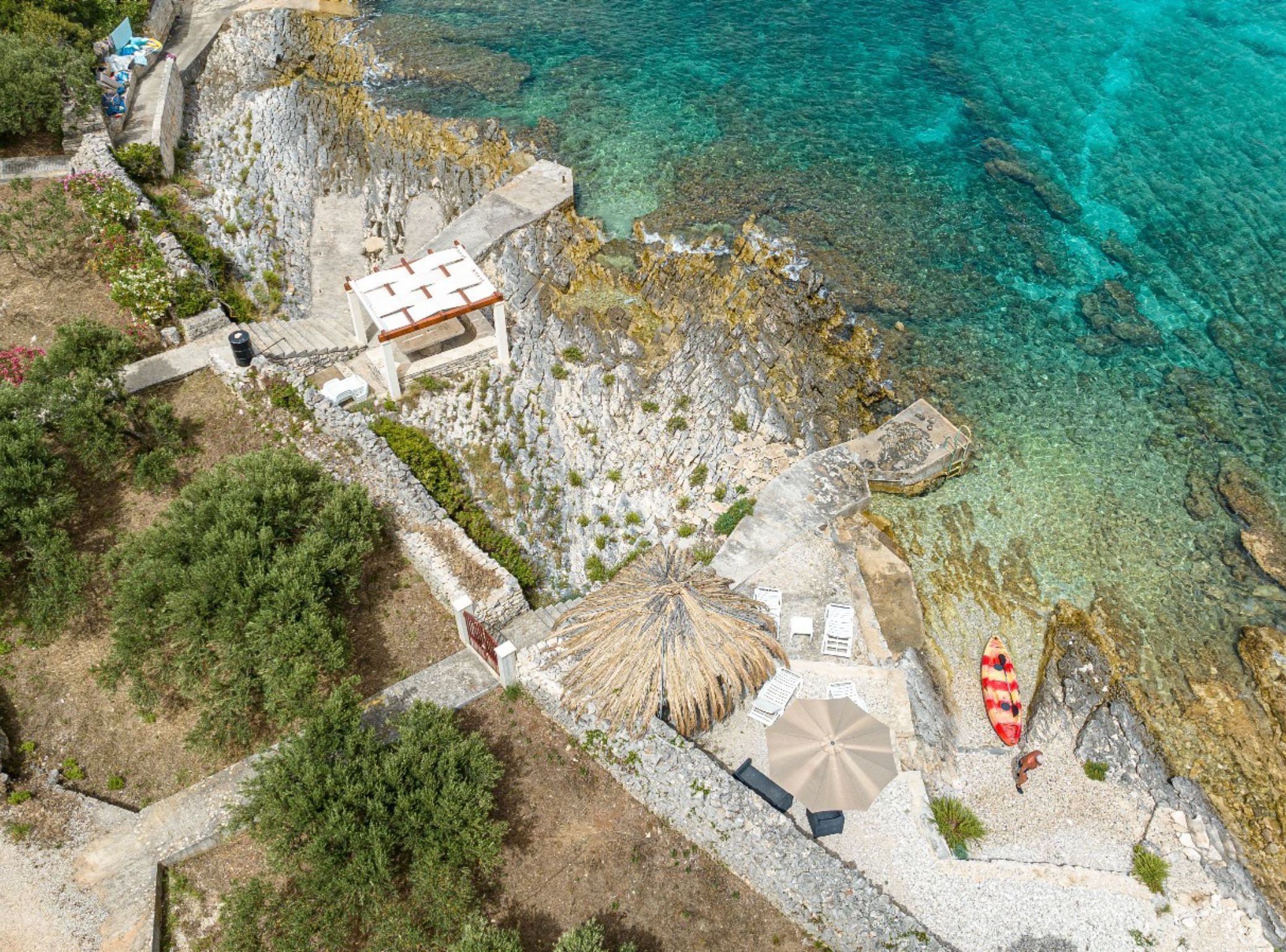 An aerial view of a coastal property with a stone pathway leading to a beach area with a thatched umbrella and lounge chairs.