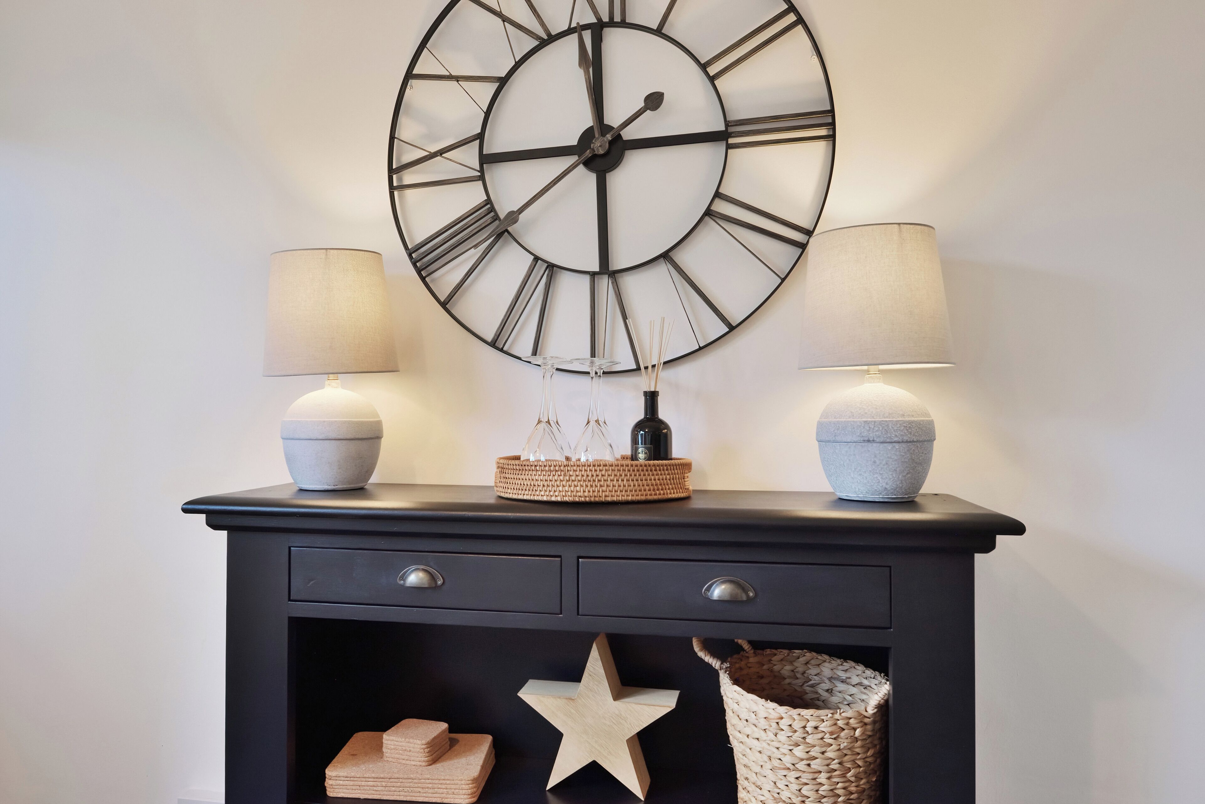 Una consola negra con dos lámparas de pared, un reloj de pared grande y objetos decorativos, incluyendo una estrella y cestas tejidas.