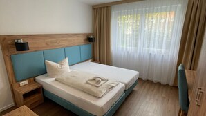 Quarto de hotel moderno com cabeceira de madeira, cama com detalhe azul, mesa de cabeceira, criado-mudo de madeira, cadeira de madeira e uma janela grande com cortinas translúcidas.