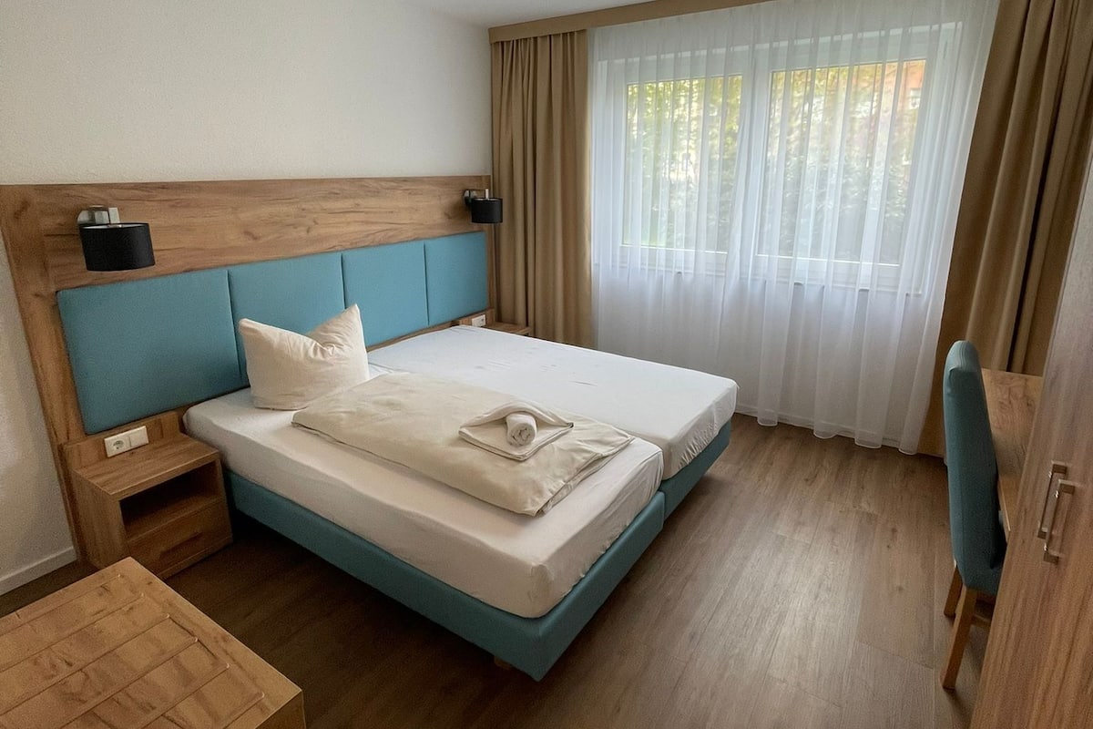 Una habitación de hotel moderna con cabecera de madera, cama con detalles azules, mesita de noche, tocador de madera, una silla de madera y un ventanal con cortinas traslúcidas.