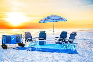 Un coin aménagé sur la plage avec un chariot portant l’inscription « OCEANTRAX » et un panneau indiquant « Équipement de plage offert », comprenant des chaises, des serviettes, une glacière, un chariot, un parasol et une couverture.