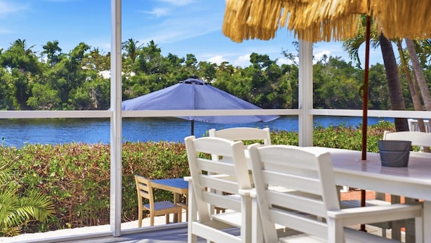 Une table entourée de chaises et surmontée d’un parasol en chaume, donnant sur une étendue d’eau, avec une végétation luxuriante en arrière-plan.