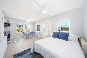 Quarto com cama, escrivaninha, cadeira, ventilador de teto e janelas com vista para áreas verdes.