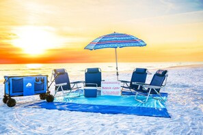 Montaje en la playa que incluye un carrito con la etiqueta "OCEANTRAX" y un letrero que indica "Equipo de playa gratuito", con sillas, toallas, hielera, carrito, sombrilla y manta.