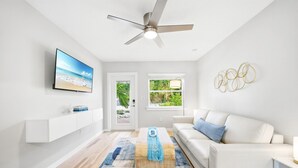 Un salon moderne doté d’un téléviseur à écran plat, d’un ventilateur de plafond, d’un canapé blanc, d’une table basse et offrant une vue sur la verdure par une fenêtre.