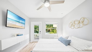 Une chambre moderne avec un grand lit, un téléviseur fixé au mur, un ventilateur de plafond et une vue sur la verdure par la fenêtre.
