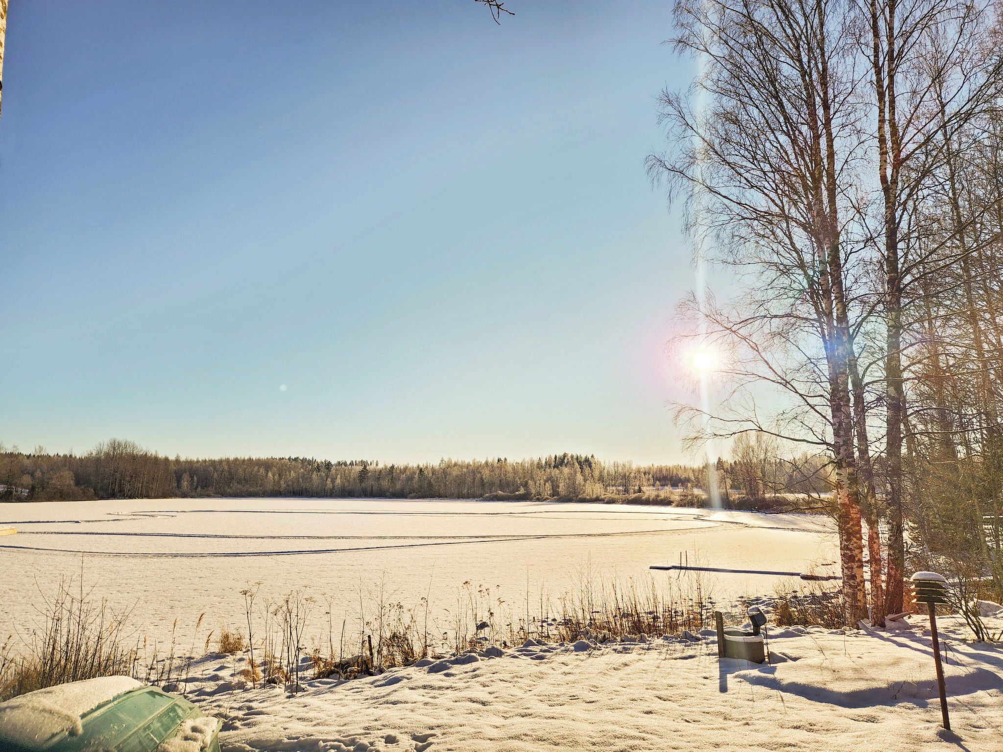 Eine verschneite Landschaft mit blauem Himmel, einem zugefrorenen Gewässer und kahlen Bäumen.
