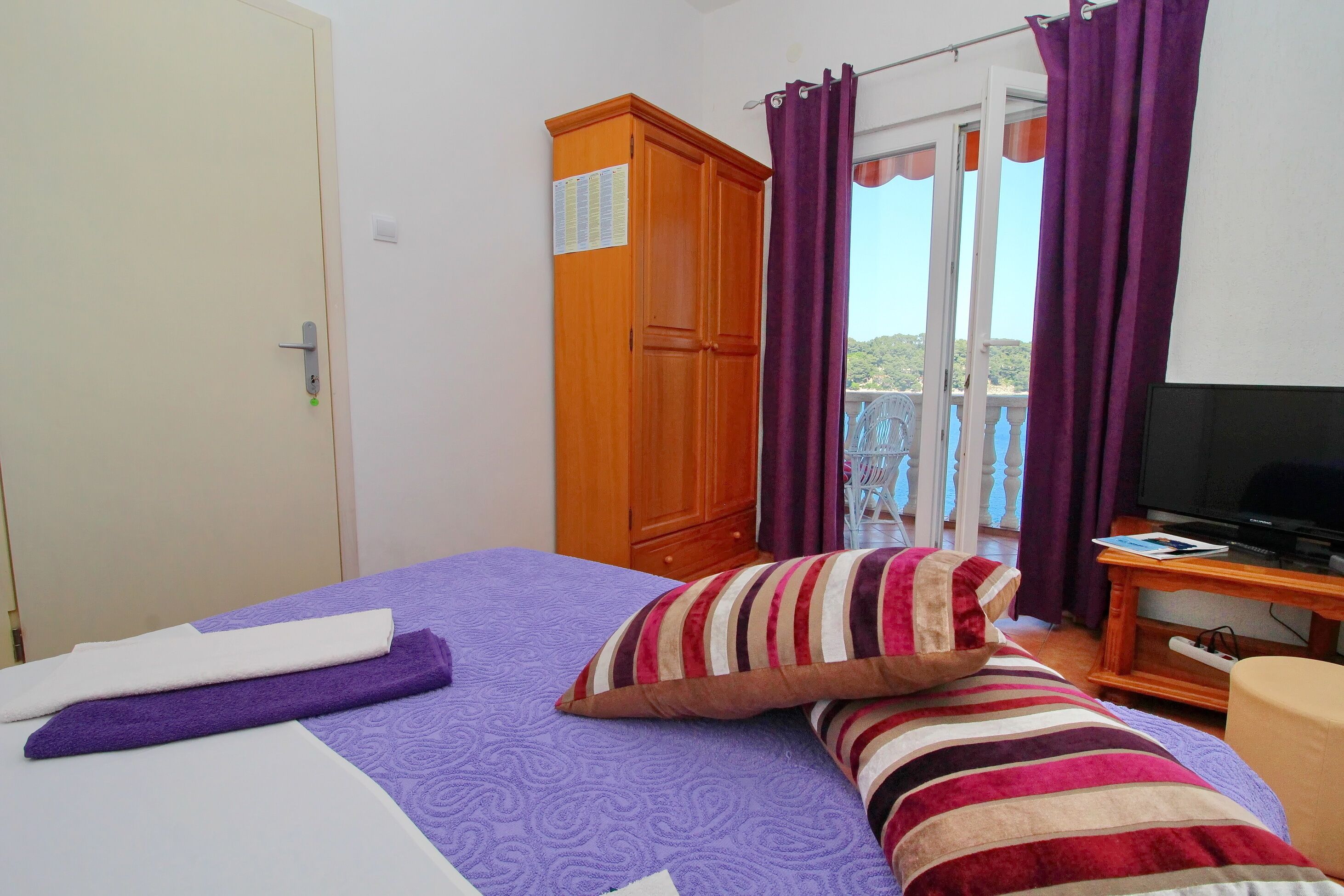 A bedroom with a bed, a wooden wardrobe, a television, and a balcony with purple curtains.