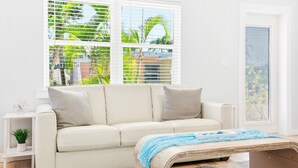 Un salon moderne avec un canapé beige, une petite table d’appoint ornée d’une plante et une fenêtre avec des stores.